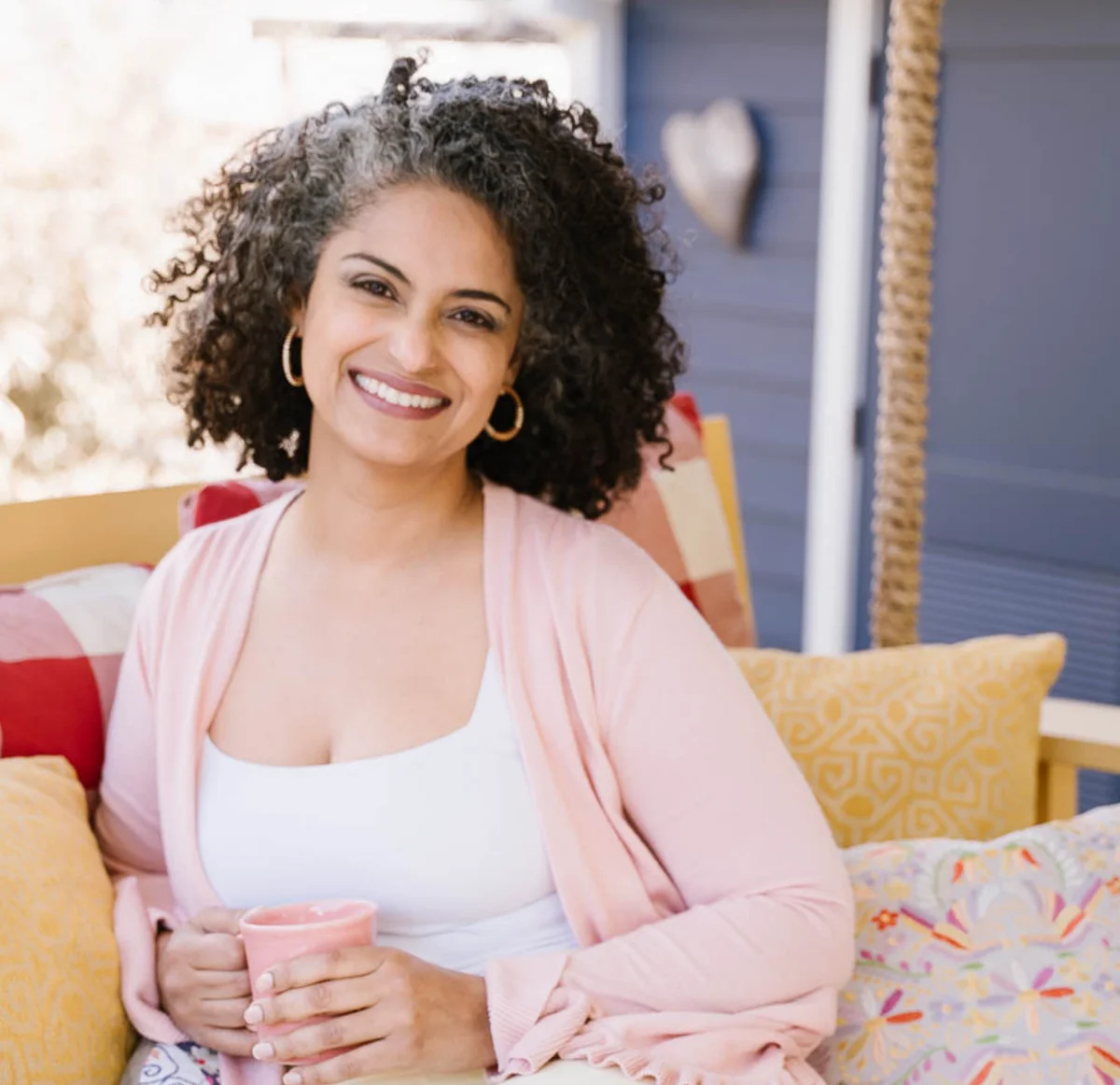 Fadya Al Bakry seated on her porch, wearing a blush cardigan and holding a pink mug, surrounded by colorful cushions